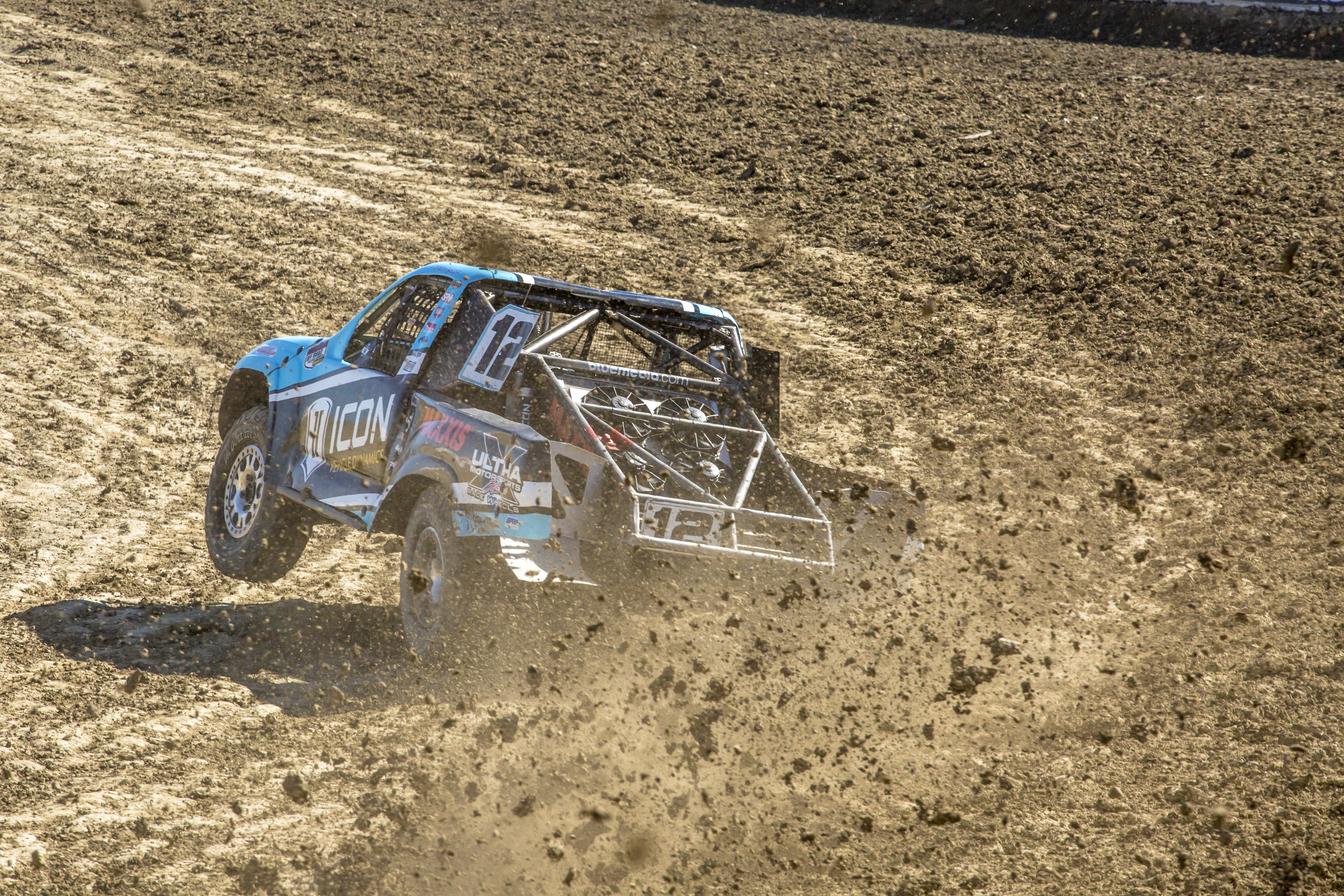 A Prolight class race truck kicking up dirt on a racetrack corner.